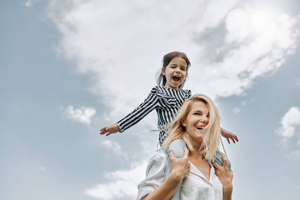 Happy little funny daughter on a piggy back ride with her happy mother on the sky background. Loving woman and her little girl playing in the park. Mom and kid has fun outside. Mother's day - 写真・画像