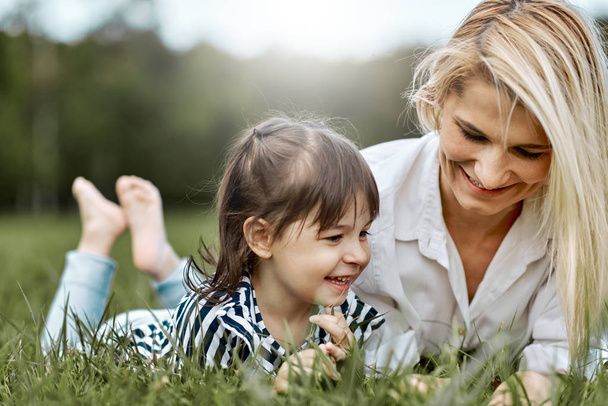 Image of happy little girl lying on the green grass and playing with her beautiful mother in the park. Woman and her little daughter enjoy the holiday. Mom and child has fun outdoors. Mother's day - Fotografie, Obrázek