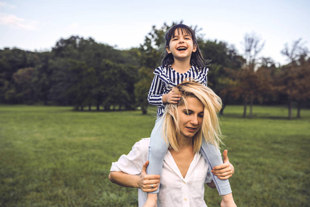 Cute happy little girl laughing and sitting on her beautiful mother's shoulders have fun outdoor in the park. Happy family time together. Positive emotion. Good relationship between mom and daughter. - Φωτογραφία, εικόνα