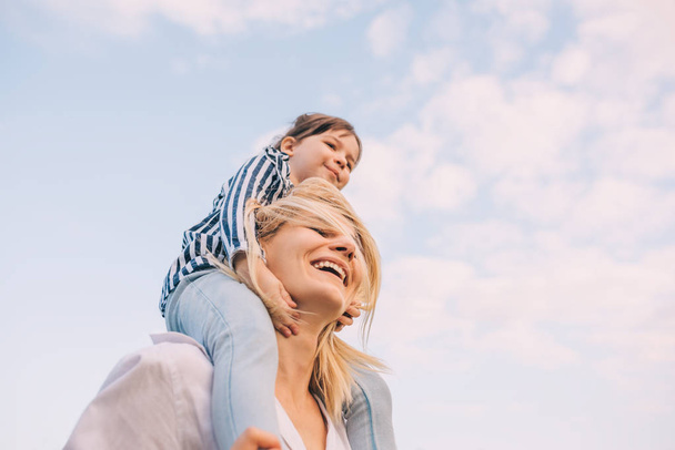 Bottom view of cute little daughter on a piggy back ride with her smiling mother on sky background. Loving woman and her little girl playing in the park. Mom and kid have fun outside. Motherhood - Φωτογραφία, εικόνα