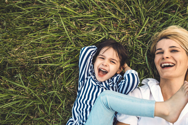 Overhead cropped image of happy beautiful woman laughing and playing with her cute little girl on green grass outdoor. Loving mother and daughter spend time together in the park. Mom and kid have fun - Foto, Imagen