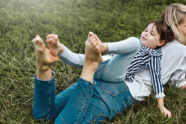 Horizontal image of beautiful woman lying and playing with her cute little girl on the green grass outdoors. Happy child and mother spend time together in the park. Mother's day concept - 写真・画像
