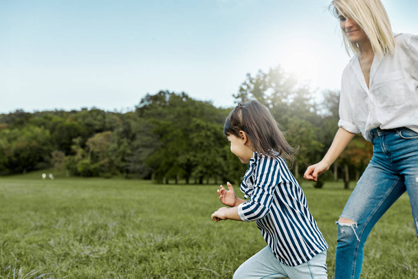 Happy pretty little girl playing catch me with her mother on green grass in the park. Loving beautiful woman and her daughter spending time together. Mom and kid has fun. Mother's day - 写真・画像