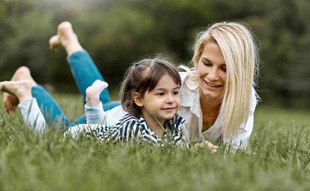 liebliches, glückliches kleines Mädchen, das auf dem grünen Gras liegt und mit seiner schönen Mutter im Park spielt. Frau und ihre kleine Tochter genießen den Urlaub. Mutter und Kind haben Spaß im Freien. Muttertag - Foto, Bild