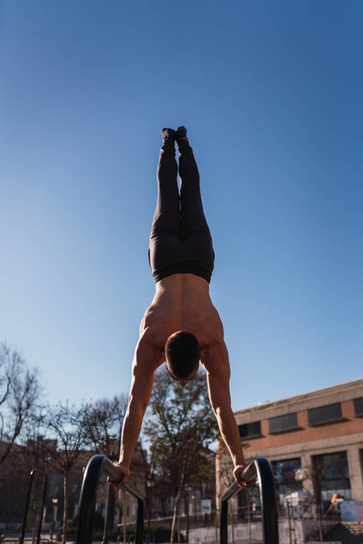 Back view of young guy doing handstand exercises on gymnastic parallel bars in sunny day - Foto, Bild