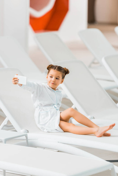 lindo niño en albornoz tomando selfie mientras está acostado en la silla de cubierta
  - Foto, Imagen