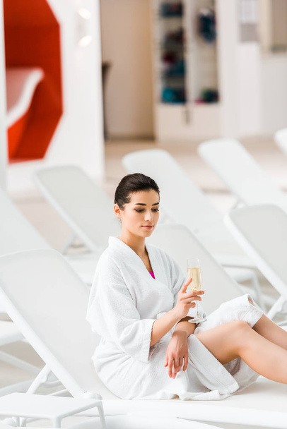 aantrekkelijke vrouw houden glas champagne en zitten in badjas op ligstoel  - Foto, afbeelding