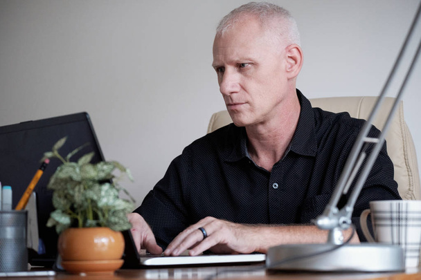Skoncentrowany siwy dojrzałe kaukaski mężczyzna siedzący przy stole office i odpowiadającego z partnerem biznesowym na laptopie - Zdjęcie, obraz