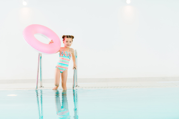 adorable kid standing in swimwear with inflatable ring and holding handrail in swimming pool - Fotó, kép