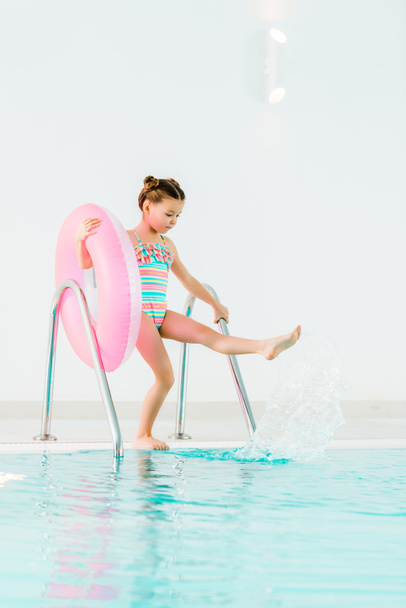 adorable kid in swimwear holding inflatable ring and playing with water in swimming pool - Photo, Image