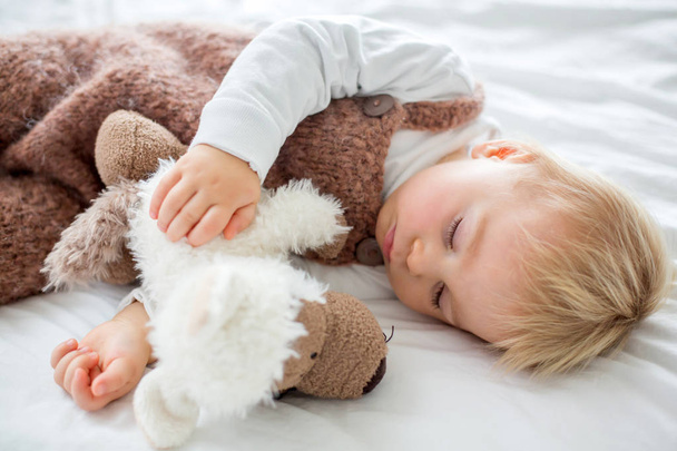 Sweet baby boy in cute overall, sleeping in bed with teddy bear stuffed toys, winter landscape behind him - Foto, afbeelding