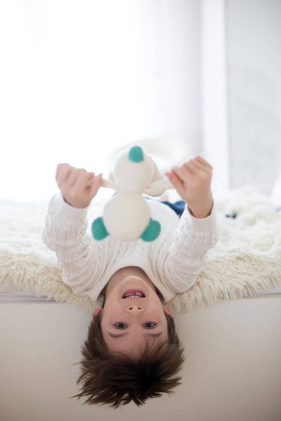Cute portrait of child upside down, lying on bed, smiling at camera and playing with toy - Photo, Image