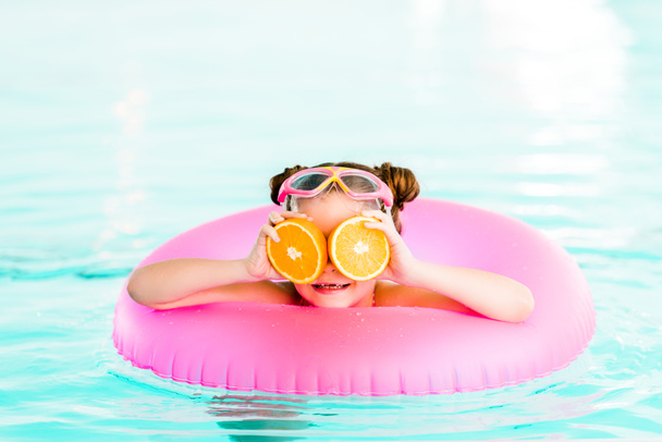 щаслива дитина тримає половину апельсинів біля очей під час плавання з надувним кільцем у басейні
 - Фото, зображення