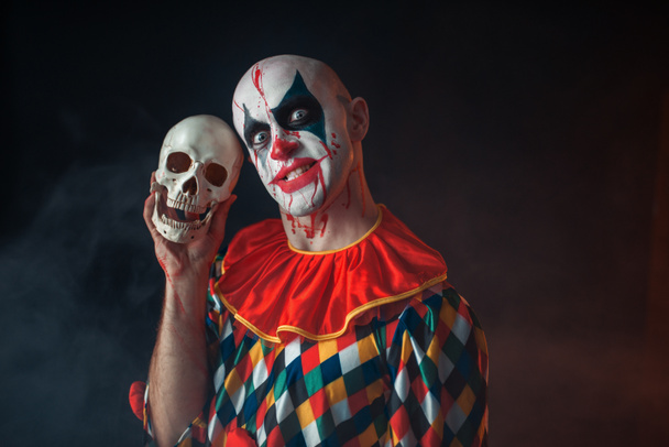 Bloody clown with crazy face holds human skull, terrible secret. Man with makeup in carnival costume, mad maniac - Photo, Image