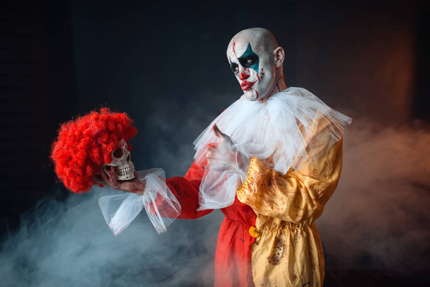 El maldito payaso loco sostiene el cráneo humano con peluca roja, horror. Hombre con maquillaje disfrazado de carnaval, loco maníaco
 - Foto, Imagen