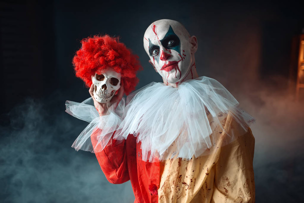 Crazy bloody clown holds human skull in red wig, horror. Man with makeup in carnival costume, mad maniac - Fotó, kép