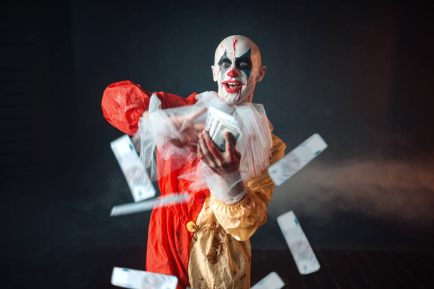 Scary bloody clown with crazy eyes holds fan of money. Man with makeup in carnival costume, mad maniac - Photo, Image
