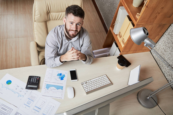 Jovem empreendedor sorridente sentado em sua mesa de escritório e olhando para a câmera, vista de cima
 - Foto, Imagem