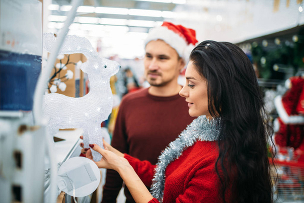 Young couple looks on christmas glass deer in supermarket, family tradition. December shopping of holiday goods and decorations - Foto, Imagen