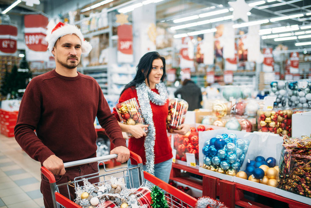 Young couple buying a lot of Christmas decorations in supermarket, family tradition. December shopping of holiday goods - Photo, Image
