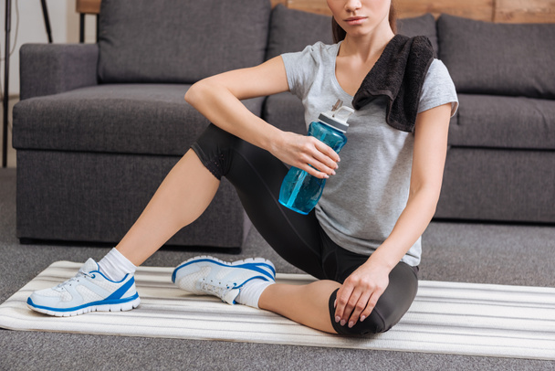 cropped view of sportswoman with towel and sport bottle sitting in living room  - Fotografie, Obrázek