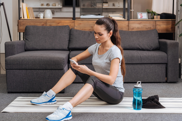 sportiva seduta su tappetino fitness e con smartphone in salotto
 - Foto, immagini