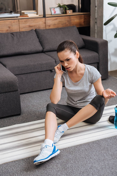 frustrated sportswoman sitting on fitness mat and talking on smartphone at home - Photo, Image