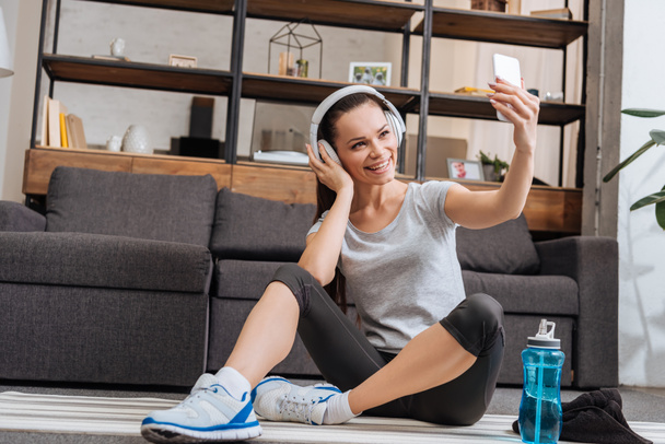 krásný usměvavý sportovkyně v sluchátka s selfie na smartphone doma - Fotografie, Obrázek