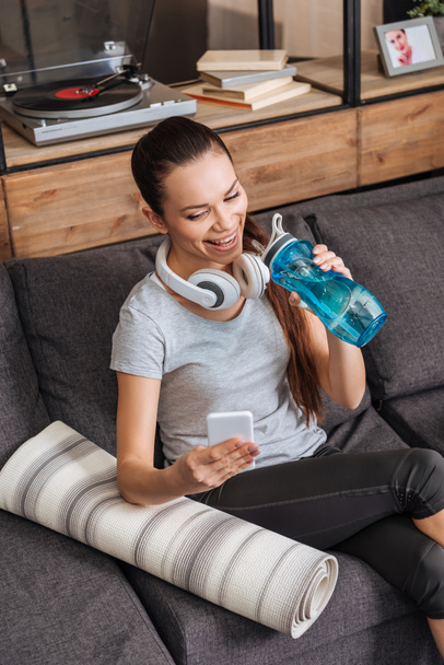 attractive sportswoman with headphones sitting on couch and using smartphone at home - Photo, Image