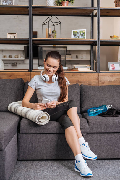 atrakcyjne sportsmenka uśmiechający się ze słuchawkami, siedząc na kanapie i w domu przy użyciu smartfona - Zdjęcie, obraz
