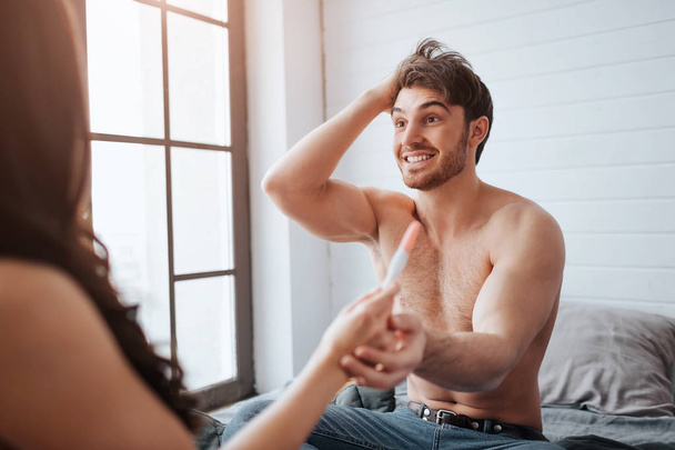 Happy young man smiling to woman. They sit on bed in room at window. Guy cheering. He hold womans hand with positive pregnancy test result. - 写真・画像