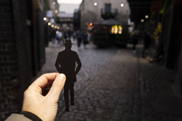 κινηματογράφηση σε πρώτο πλάνο από την πλευρά ενός ανθρώπου που κρατώντας ένα χαρτί cutout στο σχήμα ενός άνδρα, φορώντας ένα καπέλο σφαιριστής, περπάτημα από την παλιά δρομάκια της πόλης, στο Λονδίνο, Ηνωμένο Βασίλειο, το σούρουπο - Φωτογραφία, εικόνα