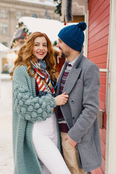 a young beautiful couple in beautiful winter clothes, a man in a gray coat, a woman in  cardigan, are standing outside in the winter near red shops - Fotó, kép