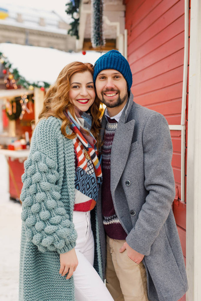 a young beautiful couple in beautiful winter clothes, a man in a gray coat, a woman in  cardigan, are standing outside in the winter near red shops - Foto, afbeelding