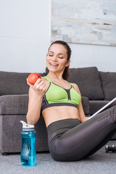beautiful sportswoman holding apple and smiling at home - Foto, Imagem
