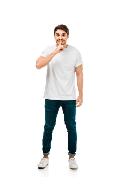 full length view of man in white t-shirt gesturing for silence with finger on lips and looking at camera isolated on white  - Foto, Imagen