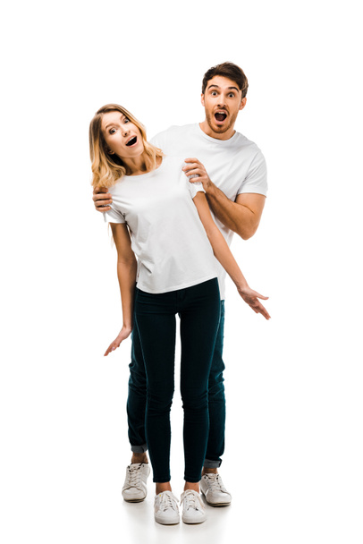Ganzkörperansicht eines überraschten jungen Paares in weißen T-Shirts, das isoliert auf weiß in die Kamera blickt  - Foto, Bild