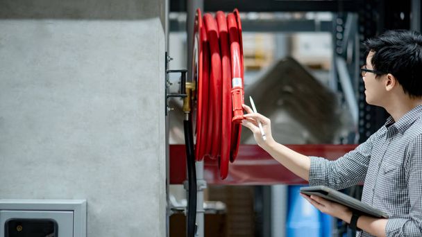 Молодой азиатский техник проверяет катушку красного пожарного шланга с помощью цифрового планшета и ручки. Концепции обслуживания зданий
 - Фото, изображение