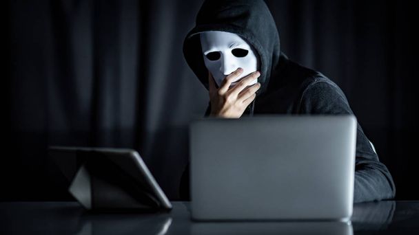 Mystery male hoodie hacker holding white mask looking at laptop computer. Anonymous social masking. Ransomware cyber attack or internet security concepts - Zdjęcie, obraz