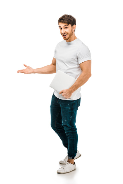 full length view of handsome young man holding laptop and smiling at camera isolated on white - Valokuva, kuva