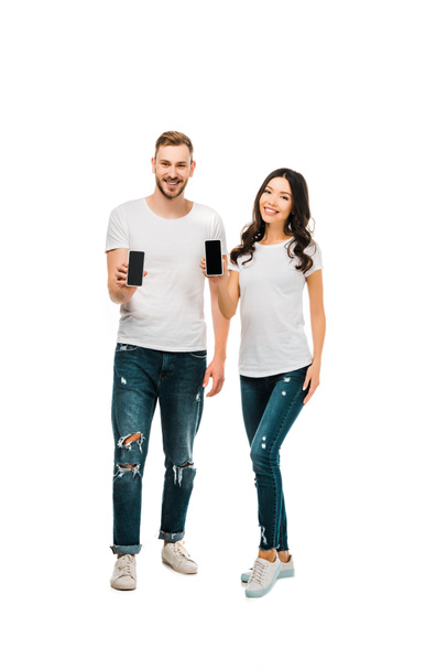 feliz joven pareja sosteniendo teléfonos inteligentes con pantallas en blanco y sonriendo a la cámara aislada en blanco
 - Foto, Imagen