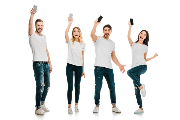 Ganzkörperansicht glücklicher junger Männer und Frauen, die Smartphones in der Hand halten und isoliert auf weiß in die Kamera lächeln - Foto, Bild