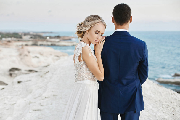 Beautiful young blonde model girl in fashionable white lace dress lean on the handsome man in the stylish blue suit and posing on the white rock at the coast of the Adriatic sea - Photo, Image