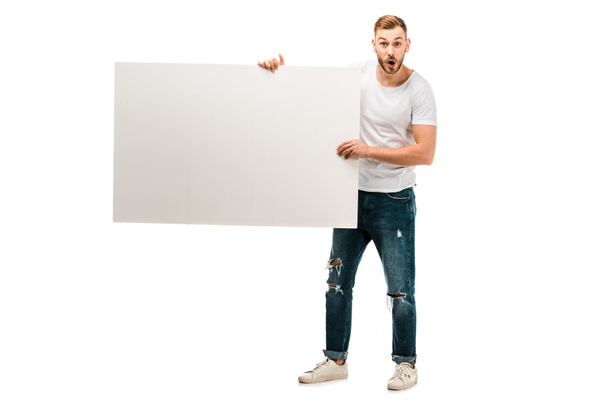shocked young man holding blank placard and looking at camera isolated on white - Foto, immagini