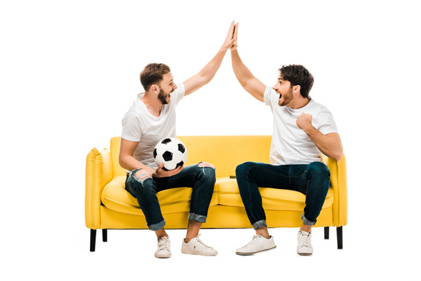 happy young man sitting on couch with soccer ball and giving high five isolated on white - Photo, Image