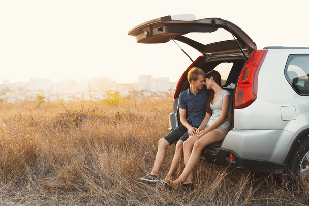 Cute young couple in love sitting in a car with an open trunk hugging ready to kiss for the first time with field in background, city on the horizon dissolve in sunlight. Travel and love concept - Photo, Image