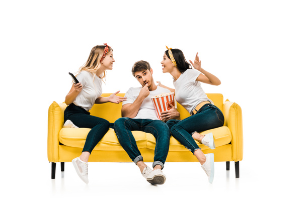 smiling young women talking and man eating popcorn while sitting together on sofa isolated on white - Fotoğraf, Görsel