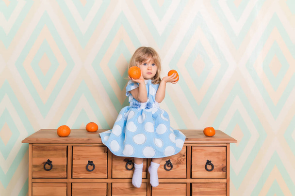 Toddler child baby girl in light blue dress seriously sitting on the wooden chest of drawers and holding fresh orange mandarins in her hands on rhomb wallpaper background. Winter holidays or christmas - Фото, изображение