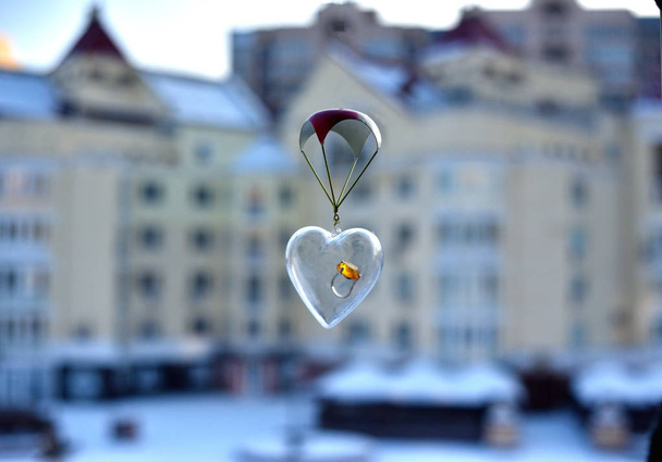 corazón transparente con un gran anillo de oro en el interior, desciende en un pequeño paracaídas en la ventana abierta sobre el fondo de las casas urbanas
. - Foto, Imagen
