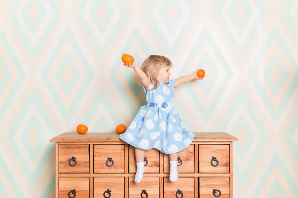 Little girl holding tangerines or oranges in her hands while sitting on the wooden wardrobe. Wearing light blue dress. Christmas holidays is over. Happy smiling baby raising hands up looking aside - Foto, Bild